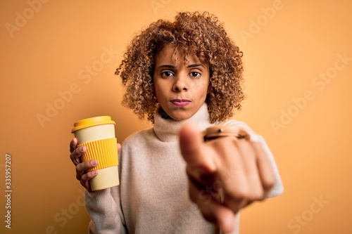 Young african american curly woman drinking cup of takeaway coffee over yellow background pointing with finger to the camera and to you, hand sign, positive and confident gesture from the front
