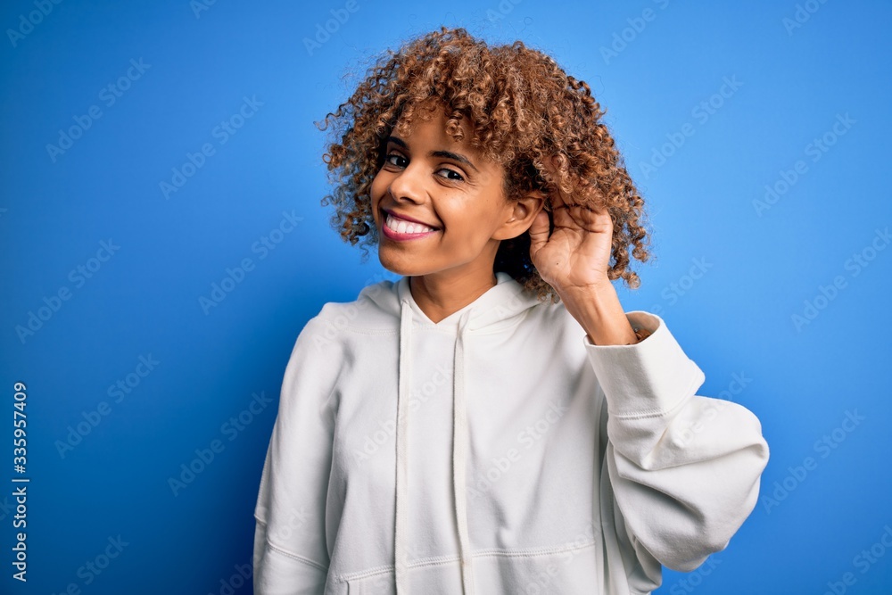 Beautiful african american sporty woman wearing casual sweatshirt over blue background smiling with hand over ear listening an hearing to rumor or gossip. Deafness concept. Beautiful african american sporty woman wearing casual sweatshirt over blue background smiling with hand over ear listening an hearing to rumor or gossip. Deafness concept.