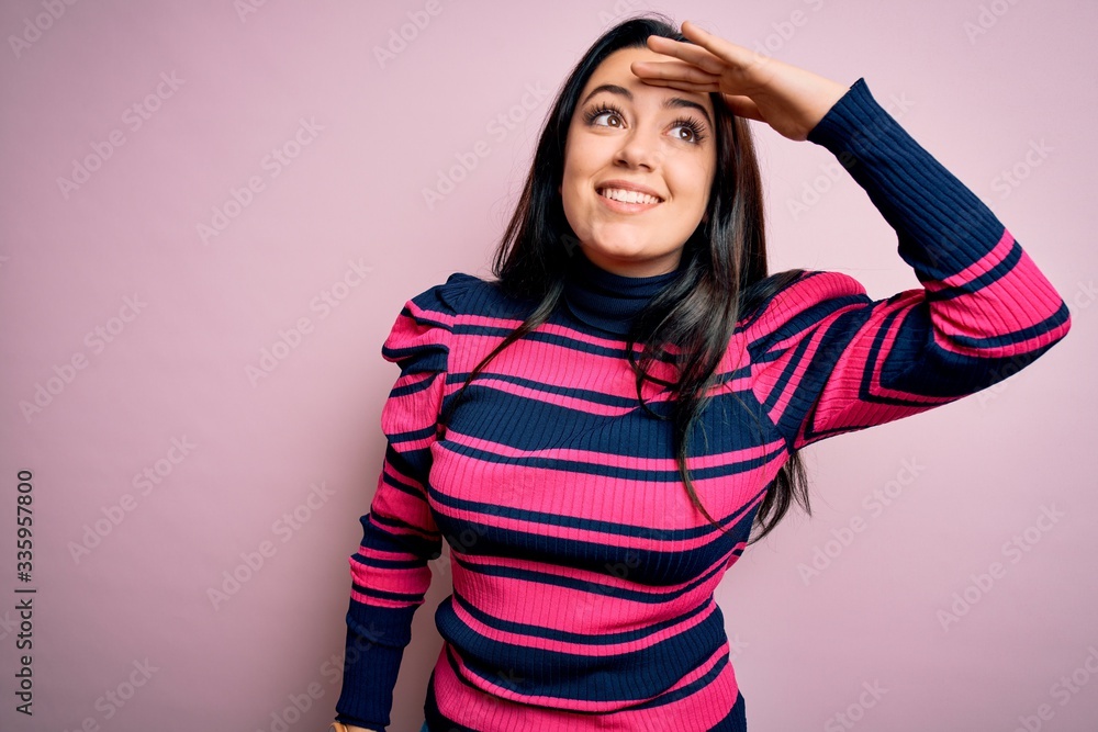 Young brunette elegant woman wearing striped shirt over pink isolated background very happy and smiling looking far away with hand over head. Searching concept. Young brunette elegant woman wearing striped shirt over pink isolated background very happy and smiling looking far away with hand over head. Searching concept.