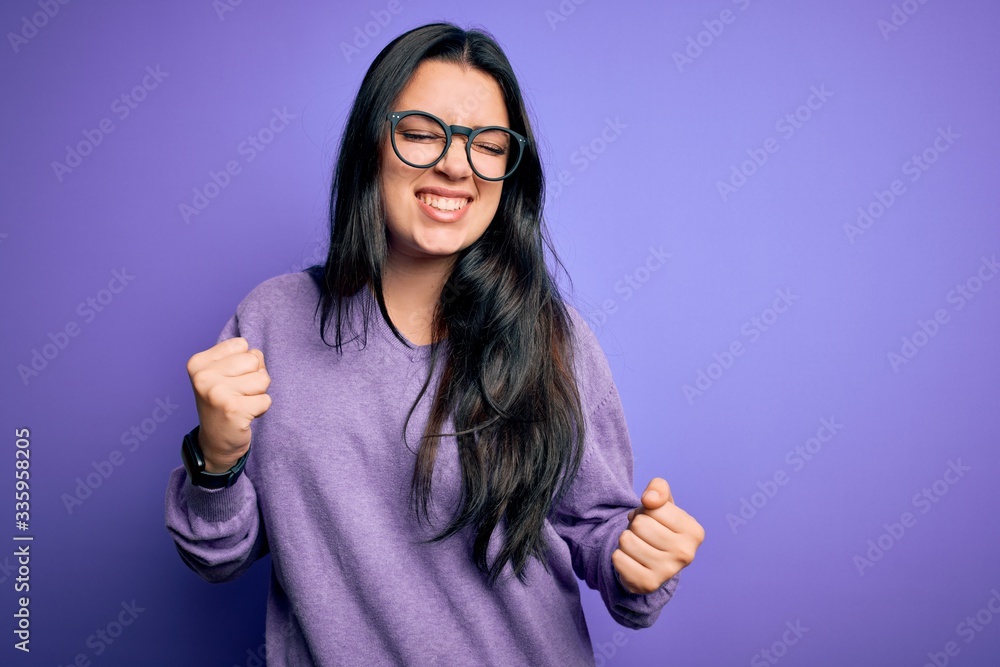 Young brunette woman wearing glasses over purple isolated background very happy and excited doing winner gesture with arms raised, smiling and screaming for success. Celebration concept. Young brunette woman wearing glasses over purple isolated background very happy and excited doing winner gesture with arms raised, smiling and screaming for success. Celebration concept.