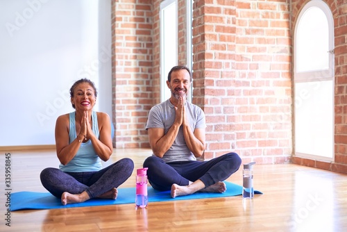 Middle age sporty couple sitting on mat doing stretching yoga exercise at gym praying with hands together asking for forgiveness smiling confident.