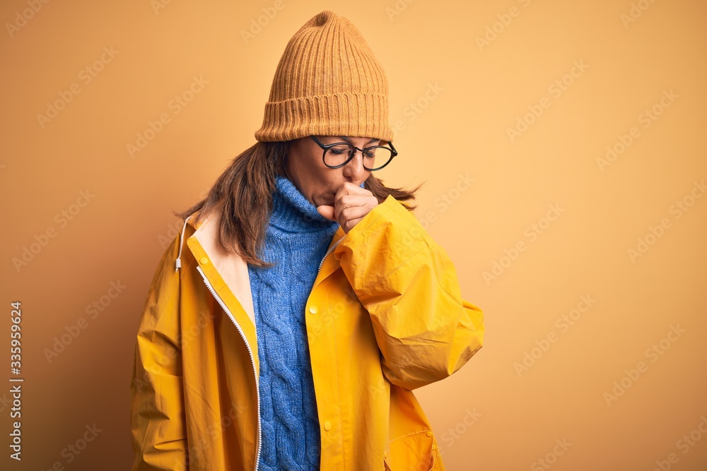 Middle age woman wearing yellow raincoat and winter hat over isolated background feeling unwell and coughing as symptom for cold or bronchitis. Health care concept. Middle age woman wearing yellow raincoat and winter hat over isolated background feeling unwell and coughing as symptom for cold or bronchitis. Health care concept.
