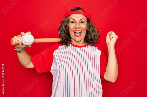 Middle age senior woman wearing baseball equiment, bat and ball over red isolated background screaming proud and celebrating victory and success very excited, cheering emotion photo