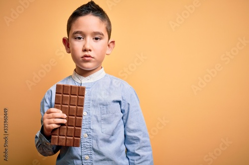 Young little boy kid eating sweet chocolate bar for dessert over isolated yellow background with a confident expression on smart face thinking serious