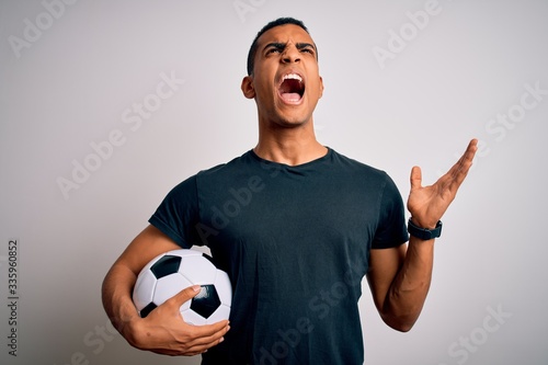Handsome african american man playing footbal holding soccer ball over white background crazy and mad shouting and yelling with aggressive expression and arms raised. Frustration concept. © Krakenimages.com