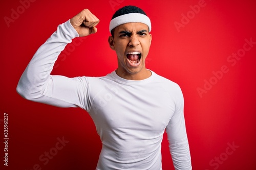 Young handsome african american sportsman wearing sportswear over red background angry and mad raising fist frustrated and furious while shouting with anger. Rage and aggressive concept.