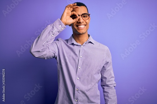Handsome african american man wearing striped shirt and glasses over purple background doing ok gesture with hand smiling, eye looking through fingers with happy face. © Krakenimages.com