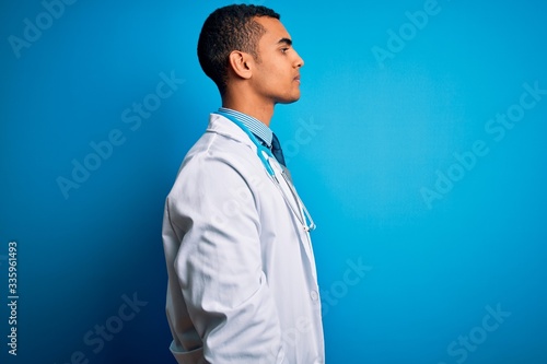 Handsome african american doctor man wearing coat and stethoscope over blue background looking to side, relax profile pose with natural face and confident smile.