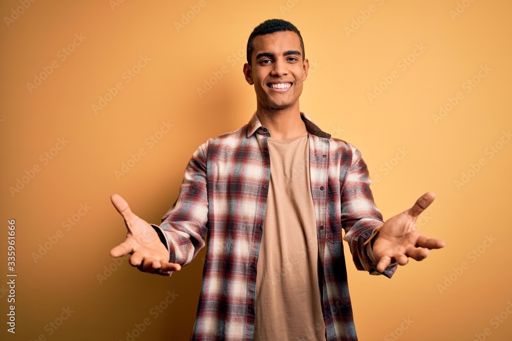 Young handsome african american man wearing casual shirt standing over yellow background smiling cheerful offering hands giving assistance and acceptance. Young handsome african american man wearing casual shirt standing over yellow background smiling cheerful offering hands giving assistance and acceptance.