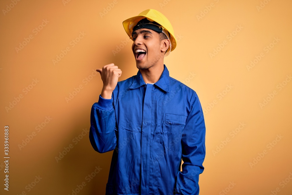 Young handsome african american worker man wearing blue uniform and security helmet smiling with happy face looking and pointing to the side with thumb up. Young handsome african american worker man wearing blue uniform and security helmet smiling with happy face looking and pointing to the side with thumb up.