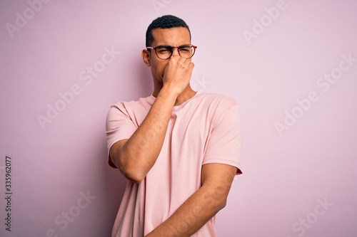 Handsome african american man wearing casual t-shirt and glasses over pink background smelling something stinky and disgusting, intolerable smell, holding breath with fingers on nose. Bad smell