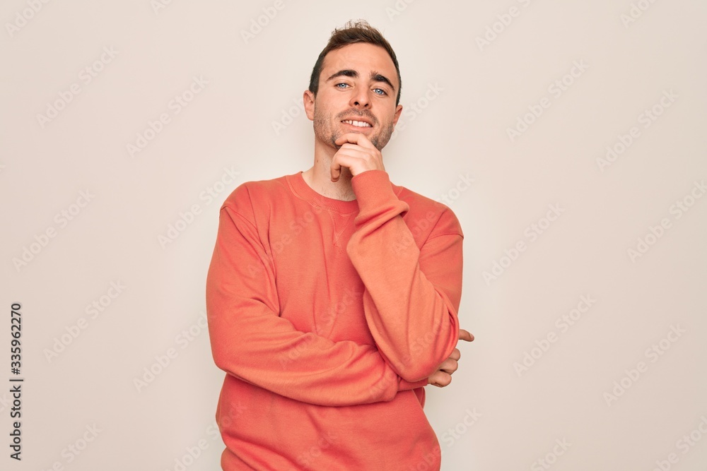 Young handsome man with blue eyes wearing casual sweater standing over white background looking confident at the camera with smile with crossed arms and hand raised on chin. Thinking positive. Young handsome man with blue eyes wearing casual sweater standing over white background looking confident at the camera with smile with crossed arms and hand raised on chin. Thinking positive.