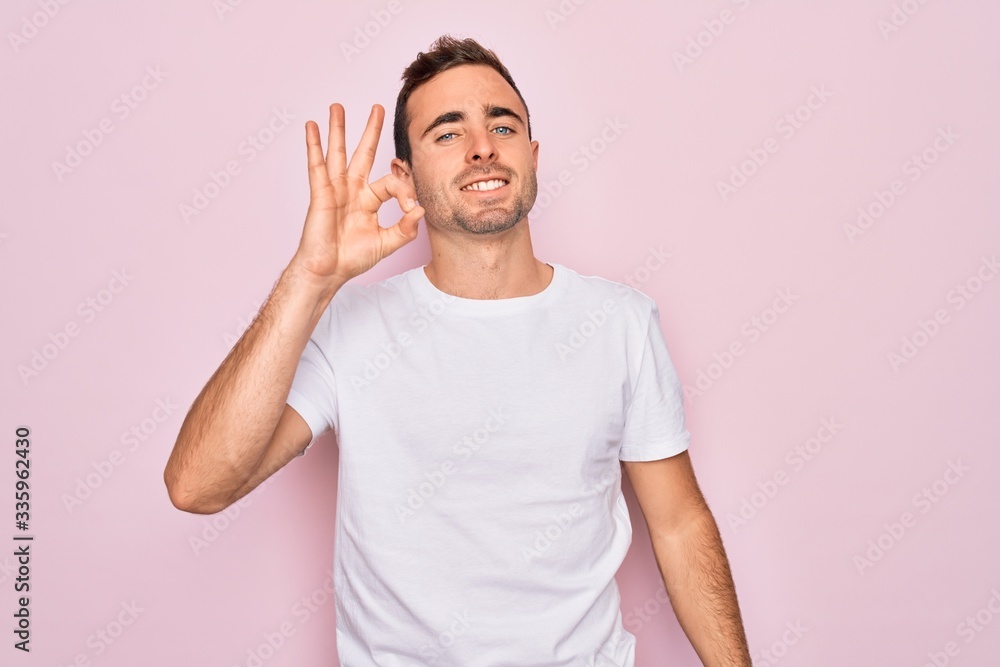 Handsome man with blue eyes wearing casual white t-shirt standing over pink background smiling positive doing ok sign with hand and fingers. Successful expression. Handsome man with blue eyes wearing casual white t-shirt standing over pink background smiling positive doing ok sign with hand and fingers. Successful expression.