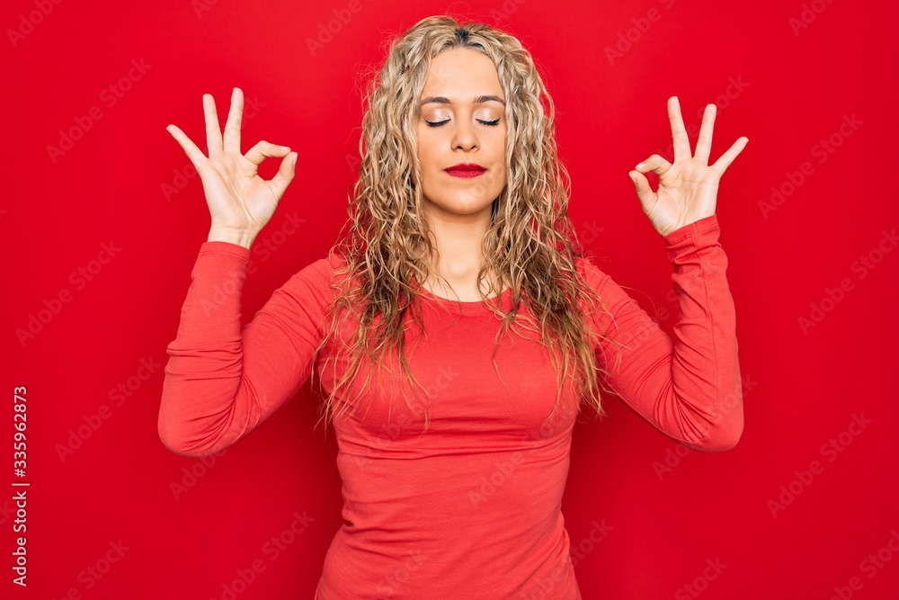 Young beautiful blonde woman wearing red casual t-shirt standing over isolated background relax and smiling with eyes closed doing meditation gesture with fingers. Yoga concept. Young beautiful blonde woman wearing red casual t-shirt standing over isolated background relax and smiling with eyes closed doing meditation gesture with fingers. Yoga concept.