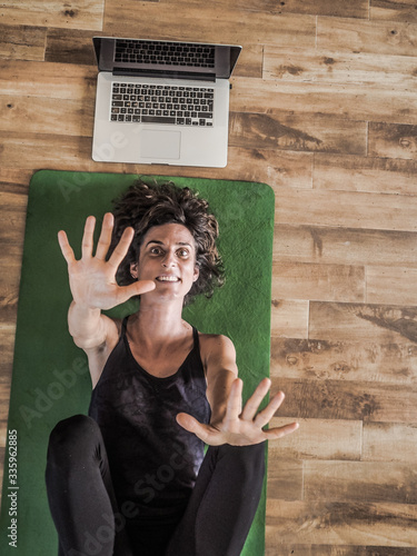 bird view of a happy smiling women lying on on a green yoga mat on wooden floor on her back with laptop open having her hands and legs into the air after yoga workout