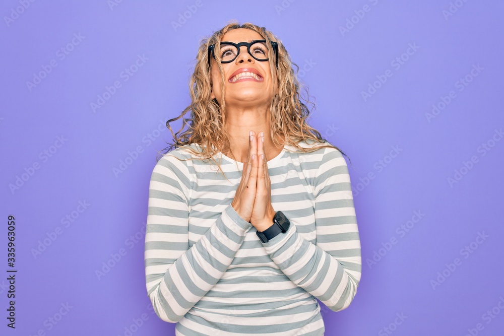 Beautiful blonde woman wearing casual striped t-shirt and glasses over purple background begging and praying with hands together with hope expression on face very emotional and worried. Begging.