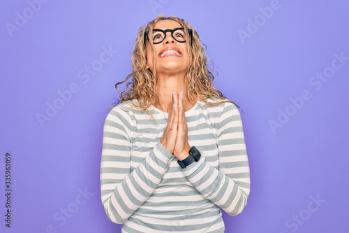 Beautiful blonde woman wearing casual striped t-shirt and glasses over purple background begging and praying with hands together with hope expression on face very emotional and worried. Begging. © Krakenimages.com