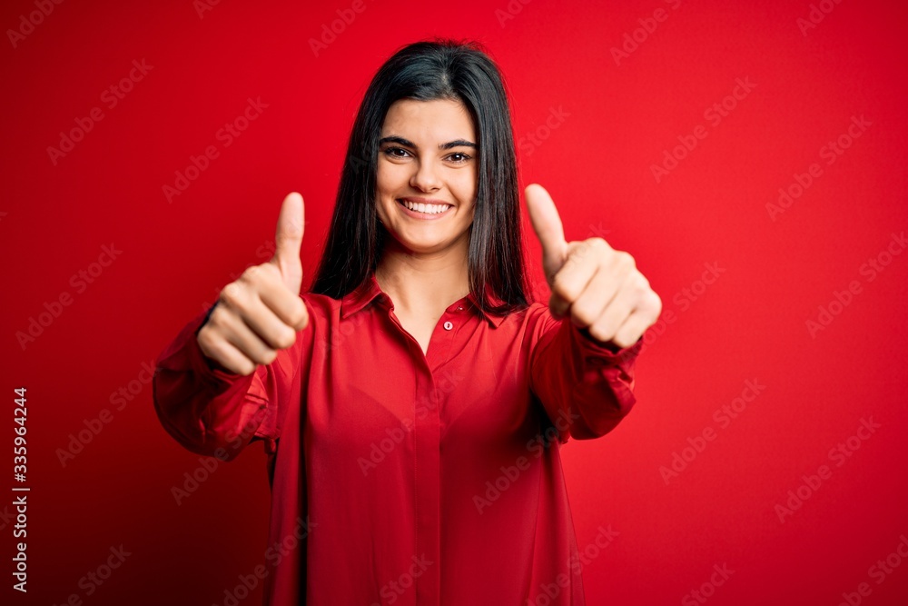 Young beautiful brunette woman wearing casual shirt standing over red background approving doing positive gesture with hand, thumbs up smiling and happy for success. Winner gesture. Young beautiful brunette woman wearing casual shirt standing over red background approving doing positive gesture with hand, thumbs up smiling and happy for success. Winner gesture.