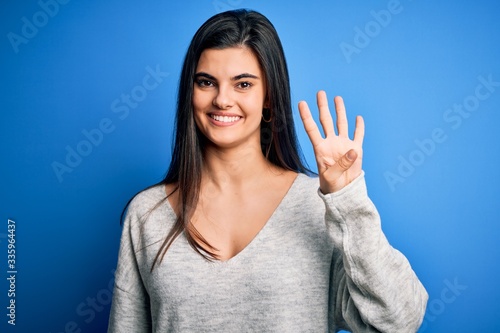 Young beautiful brunette woman wearing casual sweater standing over blue background showing and pointing up with fingers number four while smiling confident and happy.