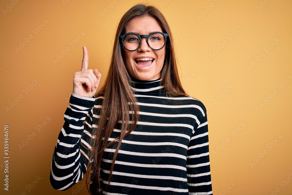 Beautiful woman with blue eyes wearing striped sweater and glasses over yellow background pointing finger up with successful idea. Exited and happy. Number one. Beautiful woman with blue eyes wearing striped sweater and glasses over yellow background pointing finger up with successful idea. Exited and happy. Number one.