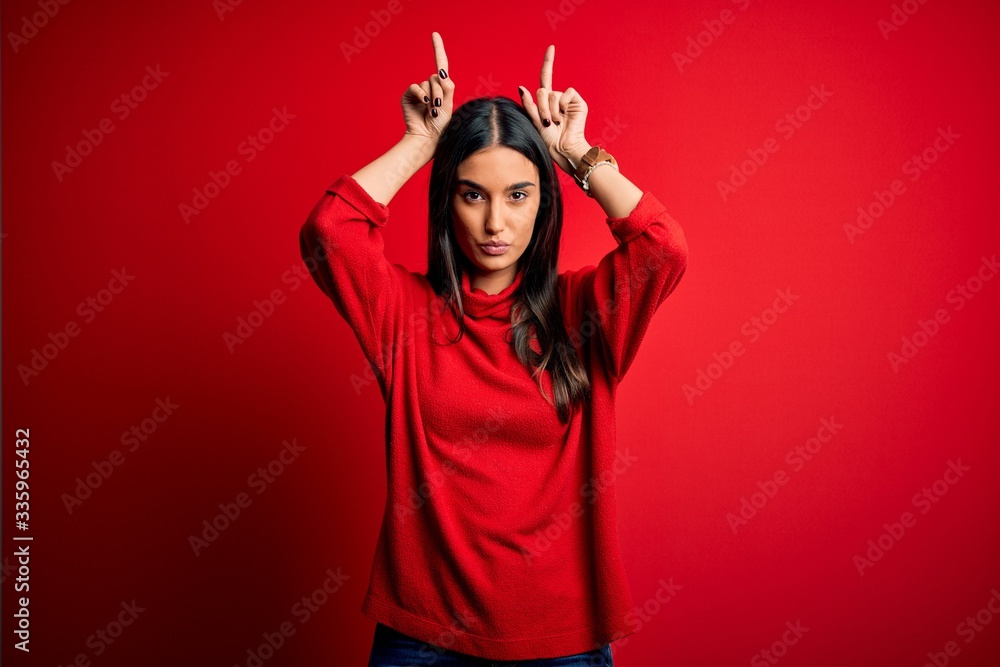 Young beautiful brunette woman wearing casual sweater over isolated red background doing funny gesture with finger over head as bull horns Young beautiful brunette woman wearing casual sweater over isolated red background doing funny gesture with finger over head as bull horns