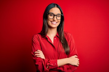 Young beautiful brunette woman wearing casual shirt and glasses over red background happy face smiling with crossed arms looking at the camera. Positive person.