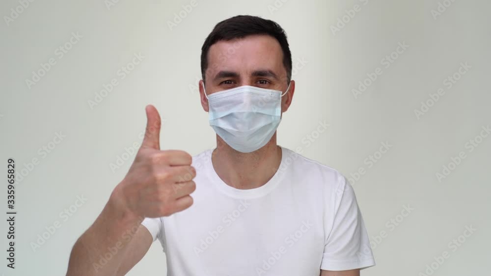 Happy Caucasian young man in medical mask is showing thumbs up gesture, close-up view of face. Concept of Coronavirus COVID-19 Pandemic. Studio shot on isolated white background. Happy Caucasian young man in medical mask is showing thumbs up gesture, close-up view of face. Concept of Coronavirus COVID-19 Pandemic. Studio shot on isolated white background.