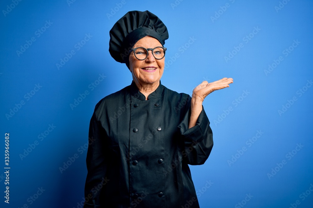 Senior beautiful grey-haired chef woman wearing cooker uniform and hat over blue background smiling cheerful presenting and pointing with palm of hand looking at the camera. Senior beautiful grey-haired chef woman wearing cooker uniform and hat over blue background smiling cheerful presenting and pointing with palm of hand looking at the camera.