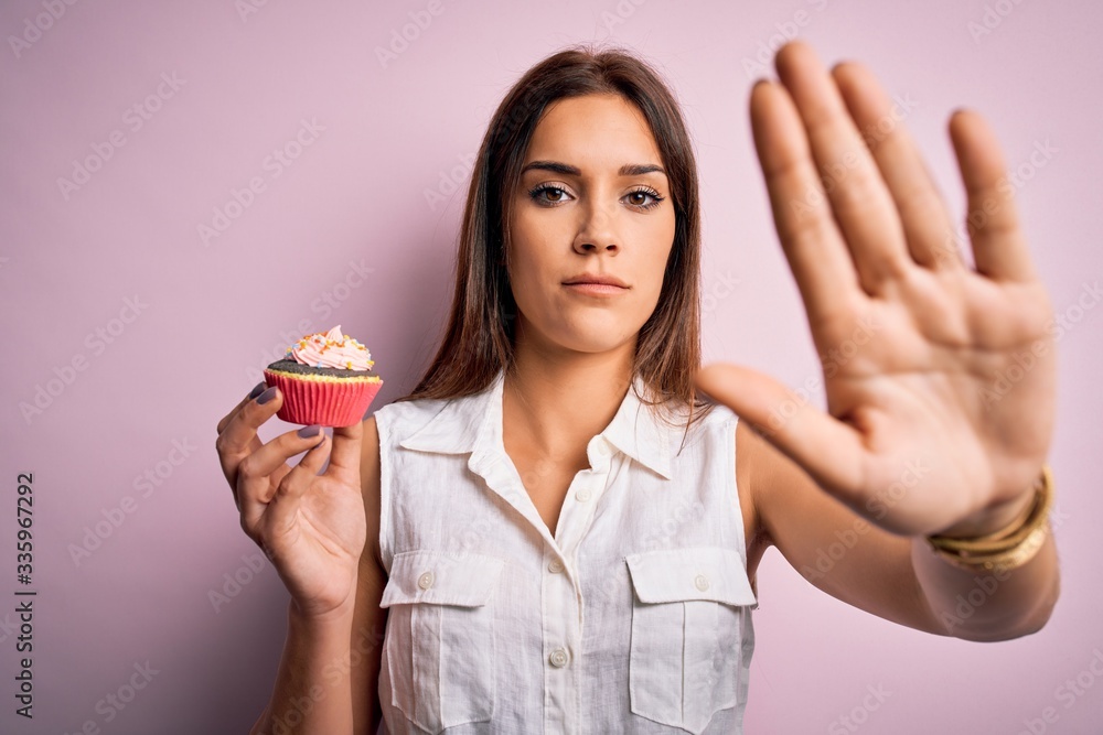Young beautiful brunette woman eating chocolate cupcake over isolated pink background with open hand doing stop sign with serious and confident expression, defense gesture Young beautiful brunette woman eating chocolate cupcake over isolated pink background with open hand doing stop sign with serious and confident expression, defense gesture