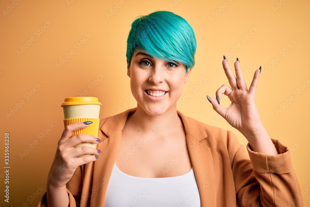 Young woman with blue fashion hair drinking a cup of coffee over yellow isolated background doing ok sign with fingers, excellent symbol Young woman with blue fashion hair drinking a cup of coffee over yellow isolated background doing ok sign with fingers, excellent symbol
