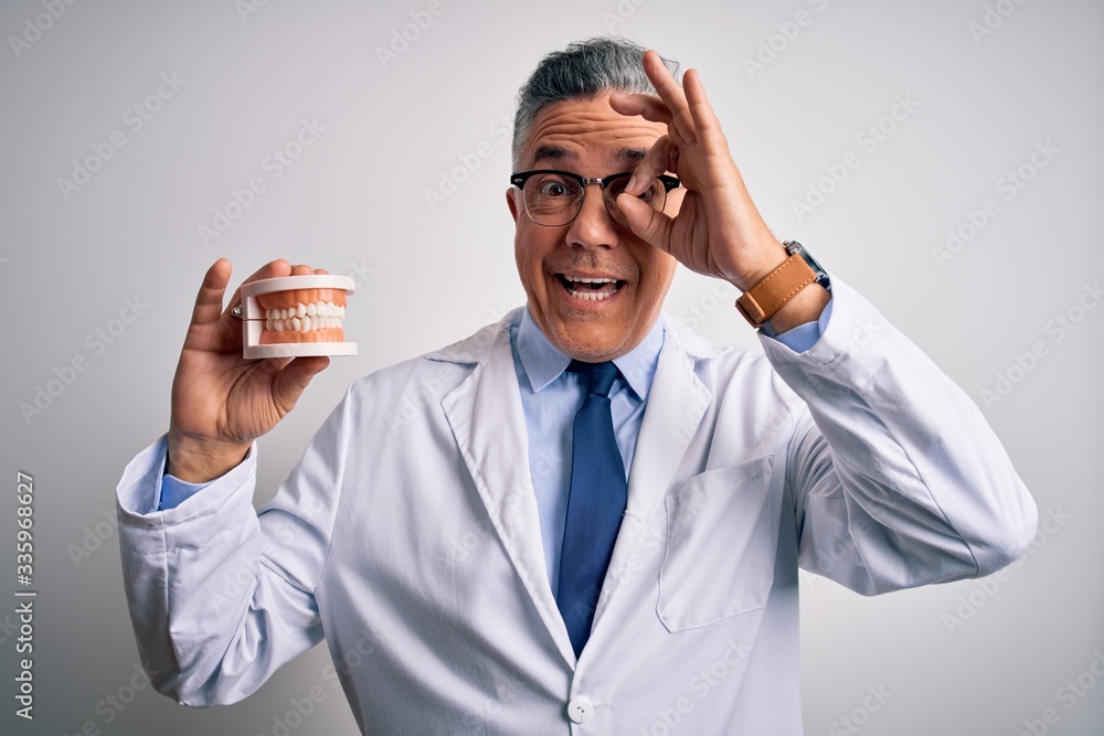 Middle age handsome grey-haired dentist man holding plastic teeth over white background with happy face smiling doing ok sign with hand on eye looking through fingers Middle age handsome grey-haired dentist man holding plastic teeth over white background with happy face smiling doing ok sign with hand on eye looking through fingers