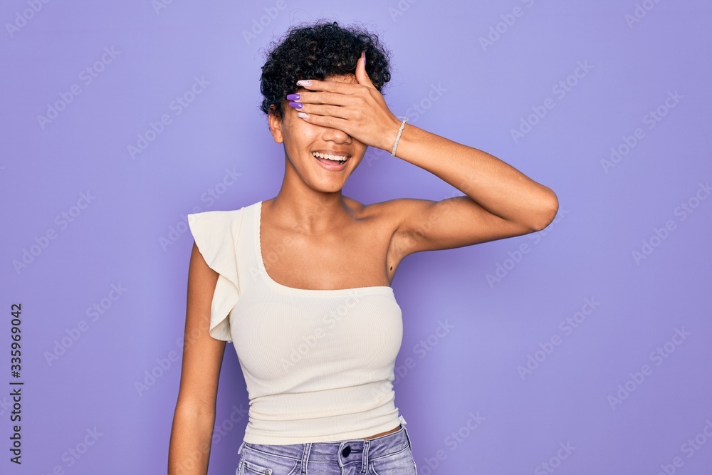 Young beautiful african american afro woman wearing casual t-shirt over purple background smiling and laughing with hand on face covering eyes for surprise. Blind concept. Young beautiful african american afro woman wearing casual t-shirt over purple background smiling and laughing with hand on face covering eyes for surprise. Blind concept.