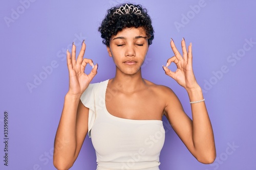 Young beautiful african american afro woman wearing tiara crown over purple background relax and smiling with eyes closed doing meditation gesture with fingers. Yoga concept.