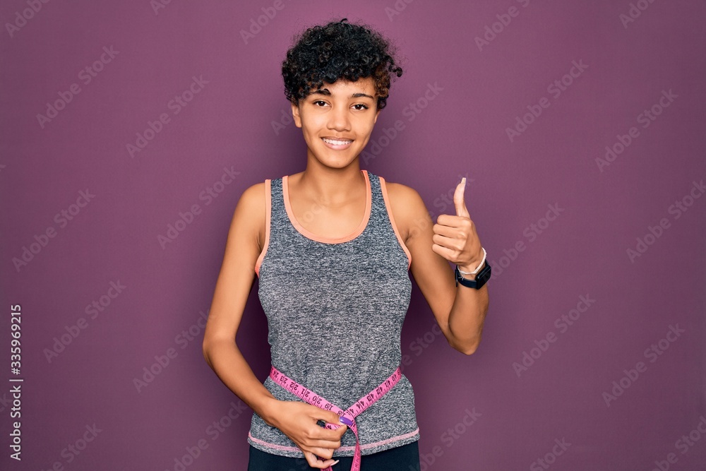 Beautiful african american sporty woman doing exercise controlling weight using tape measure happy with big smile doing ok sign, thumb up with fingers, excellent sign Beautiful african american sporty woman doing exercise controlling weight using tape measure happy with big smile doing ok sign, thumb up with fingers, excellent sign