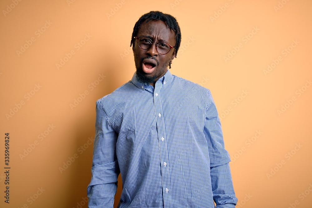 Fototapeta premium Young handsome african american man wearing shirt and glasses over yellow background In shock face, looking skeptical and sarcastic, surprised with open mouth