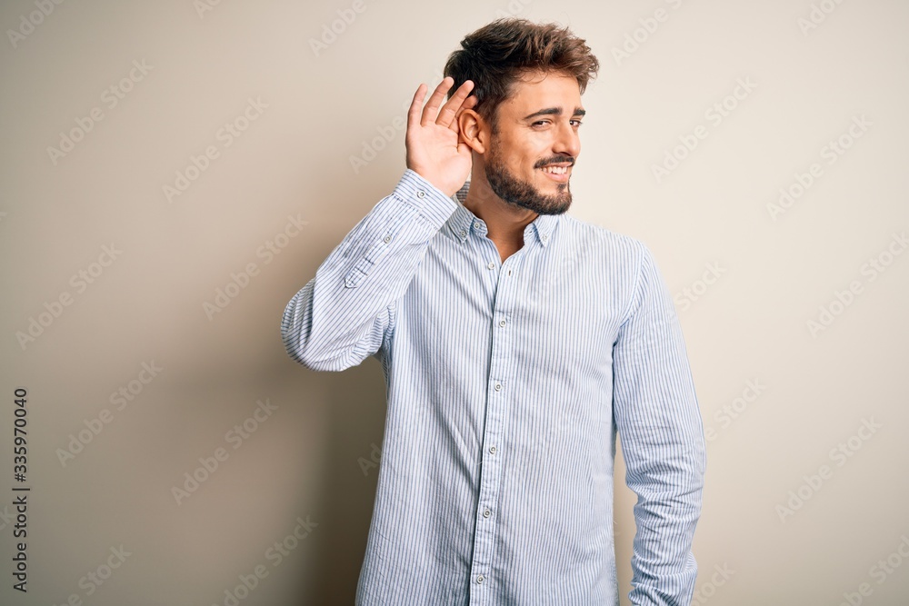 Young handsome man with beard wearing striped shirt standing over white background smiling with hand over ear listening an hearing to rumor or gossip. Deafness concept. Young handsome man with beard wearing striped shirt standing over white background smiling with hand over ear listening an hearing to rumor or gossip. Deafness concept.
