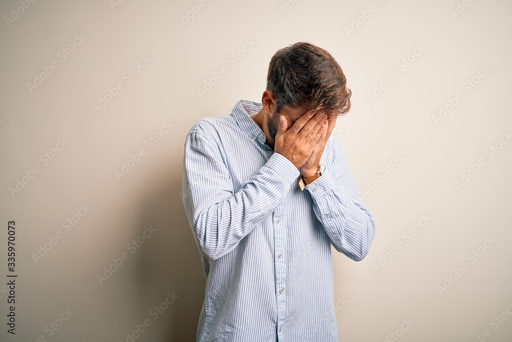 Young handsome man with beard wearing striped shirt standing over white background with sad expression covering face with hands while crying. Depression concept. Young handsome man with beard wearing striped shirt standing over white background with sad expression covering face with hands while crying. Depression concept.