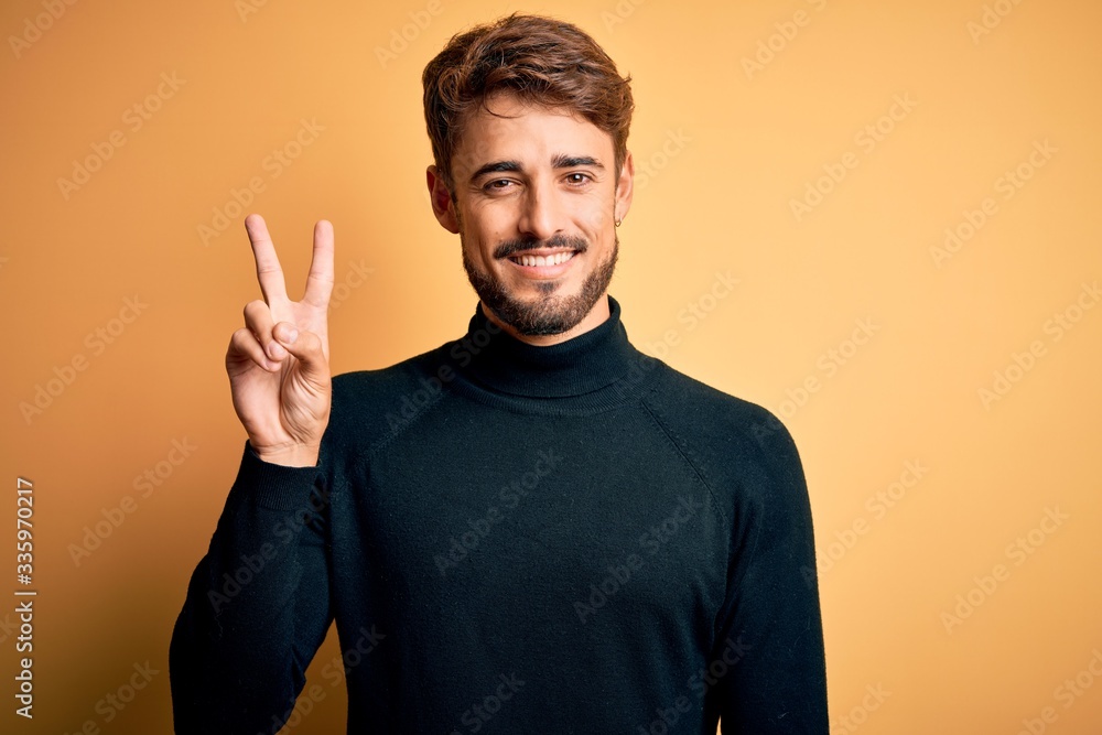 Young handsome man with beard wearing turtleneck sweater standing over yellow background showing and pointing up with fingers number two while smiling confident and happy. Young handsome man with beard wearing turtleneck sweater standing over yellow background showing and pointing up with fingers number two while smiling confident and happy.