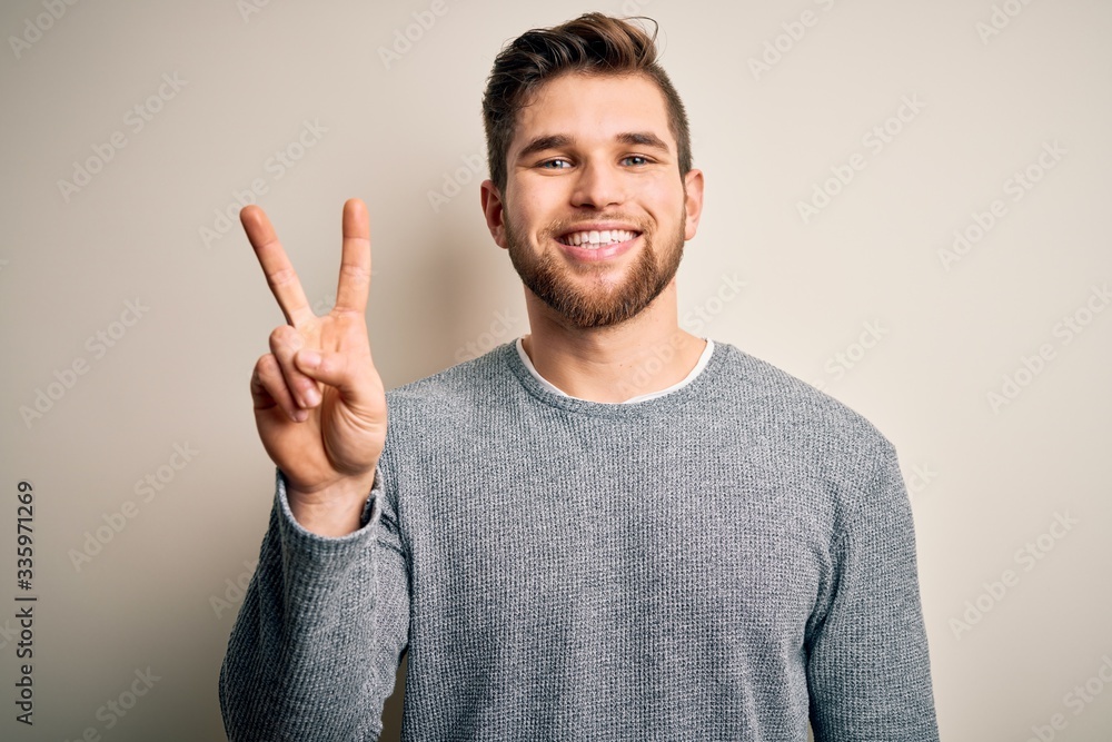Young handsome blond man with beard and blue eyes wearing casual sweater showing and pointing up with fingers number two while smiling confident and happy. Young handsome blond man with beard and blue eyes wearing casual sweater showing and pointing up with fingers number two while smiling confident and happy.