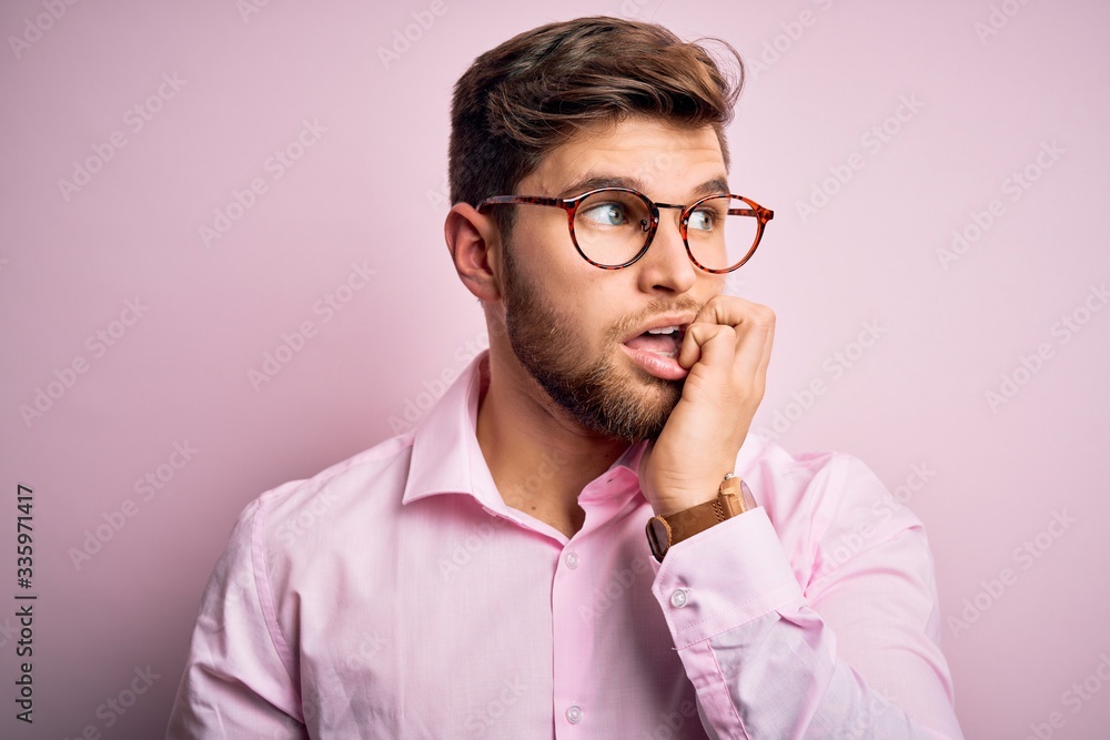 Young handsome blond man with beard and blue eyes wearing pink shirt and glasses looking stressed and nervous with hands on mouth biting nails. Anxiety problem. Young handsome blond man with beard and blue eyes wearing pink shirt and glasses looking stressed and nervous with hands on mouth biting nails. Anxiety problem.