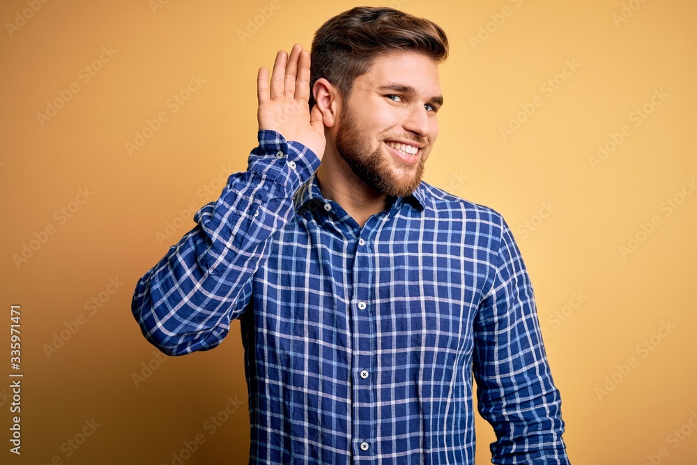 Young blond businessman with beard and blue eyes wearing shirt over yellow background smiling with hand over ear listening an hearing to rumor or gossip. Deafness concept. Young blond businessman with beard and blue eyes wearing shirt over yellow background smiling with hand over ear listening an hearing to rumor or gossip. Deafness concept.