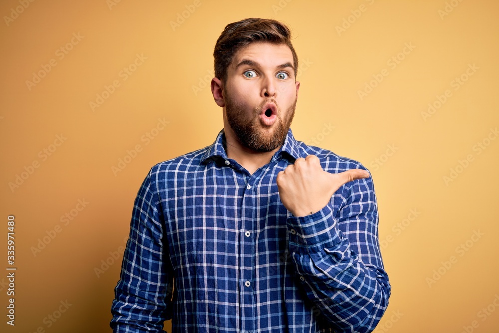 Young blond businessman with beard and blue eyes wearing shirt over yellow background Surprised pointing with hand finger to the side, open mouth amazed expression. Young blond businessman with beard and blue eyes wearing shirt over yellow background Surprised pointing with hand finger to the side, open mouth amazed expression.