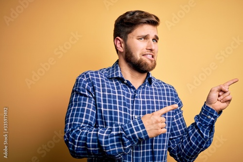 Young blond businessman with beard and blue eyes wearing shirt over yellow background Pointing aside worried and nervous with both hands, concerned and surprised expression