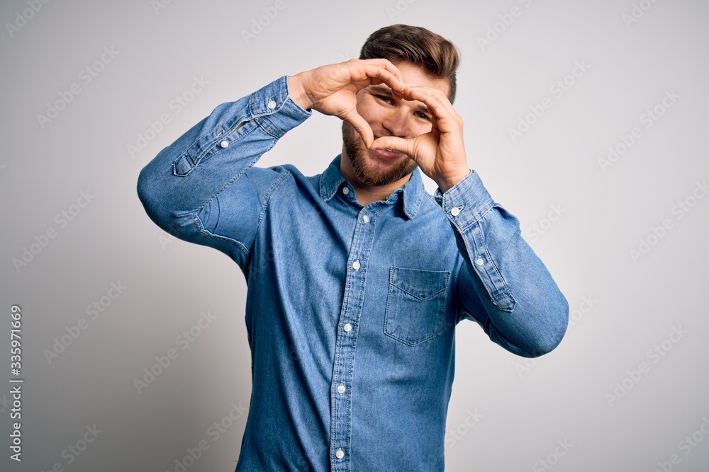 Young handsome blond man with beard and blue eyes wearing casual denim shirt Doing heart shape with hand and fingers smiling looking through sign Young handsome blond man with beard and blue eyes wearing casual denim shirt Doing heart shape with hand and fingers smiling looking through sign