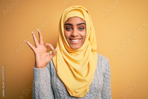 Young beautiful african american girl wearing muslim hijab over isolated yellow background smiling positive doing ok sign with hand and fingers. Successful expression.