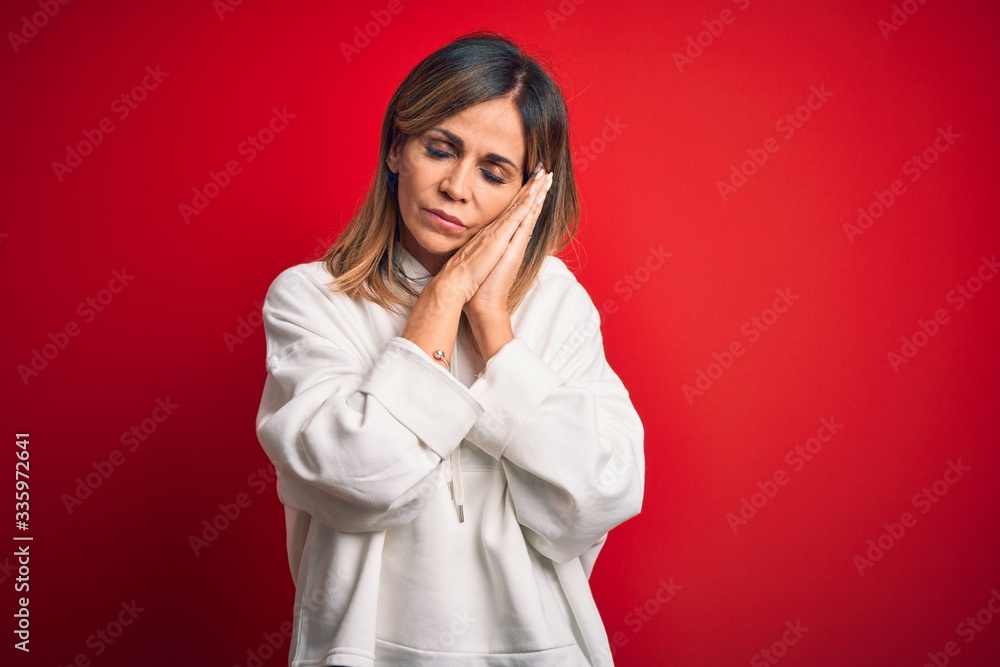 Middle age beautiful sportswoman wearing casual sweatshirt over isolated red background sleeping tired dreaming and posing with hands together while smiling with closed eyes. Middle age beautiful sportswoman wearing casual sweatshirt over isolated red background sleeping tired dreaming and posing with hands together while smiling with closed eyes.