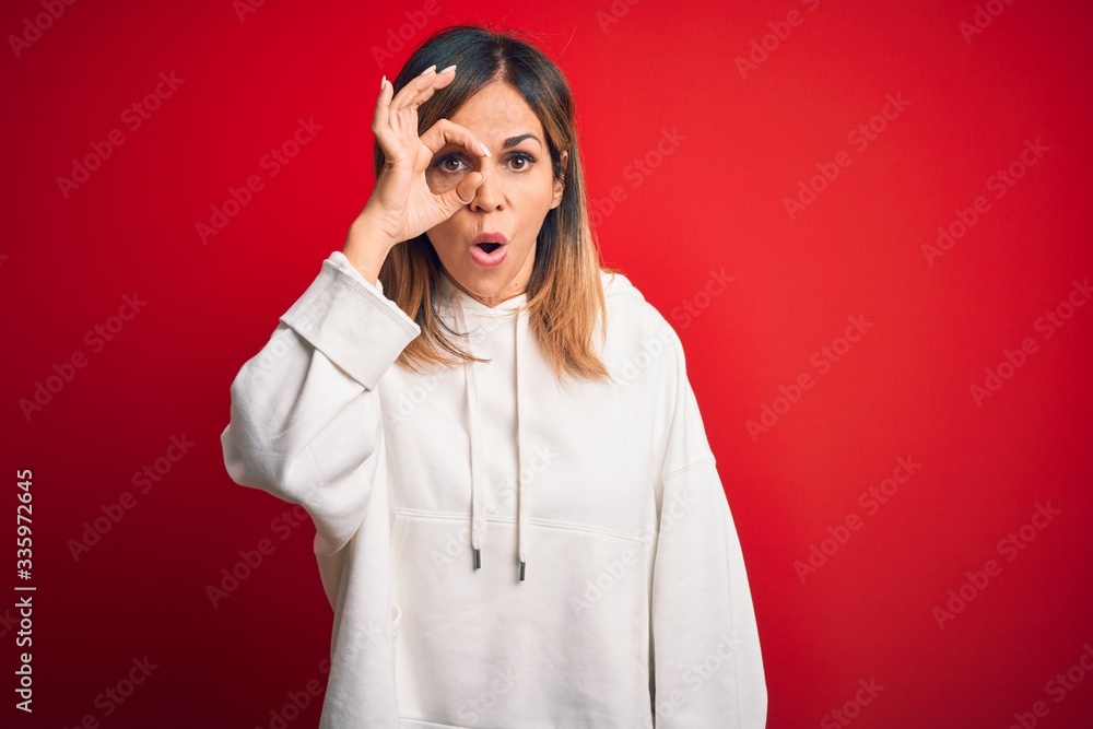Middle age beautiful sportswoman wearing casual sweatshirt over isolated red background doing ok gesture shocked with surprised face, eye looking through fingers. Unbelieving expression. Middle age beautiful sportswoman wearing casual sweatshirt over isolated red background doing ok gesture shocked with surprised face, eye looking through fingers. Unbelieving expression.