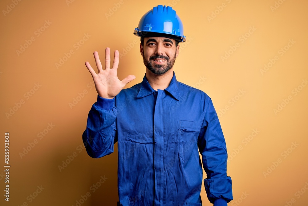 Mechanic man with beard wearing blue uniform and safety helmet over yellow background showing and pointing up with fingers number five while smiling confident and happy.