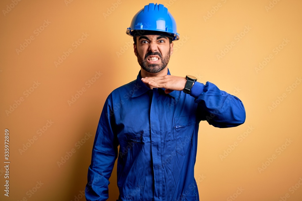 Mechanic man with beard wearing blue uniform and safety helmet over yellow background cutting throat with hand as knife, threaten aggression with furious violence