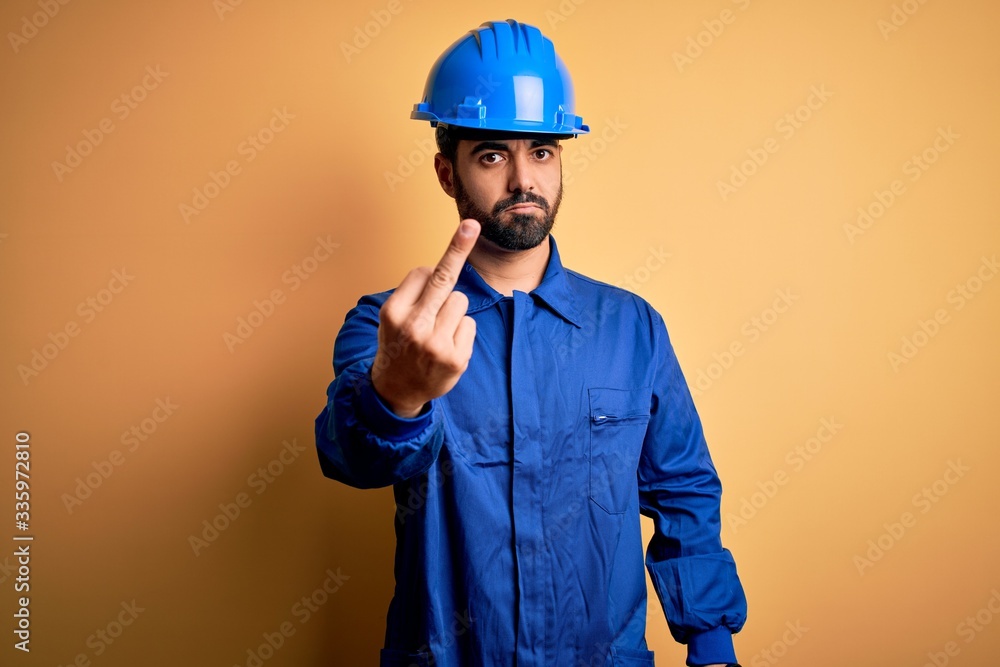 Mechanic man with beard wearing blue uniform and safety helmet over yellow background Showing middle finger, impolite and rude fuck off expression